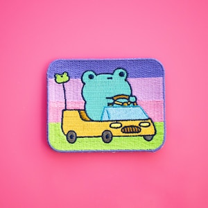 Könnte beinhalten: Ein gestickter Aufnäher mit einem grünen Frosch, der ein gelbes Auto fährt. Der Aufnäher hat einen lila, rosa und grünen Hintergrund.