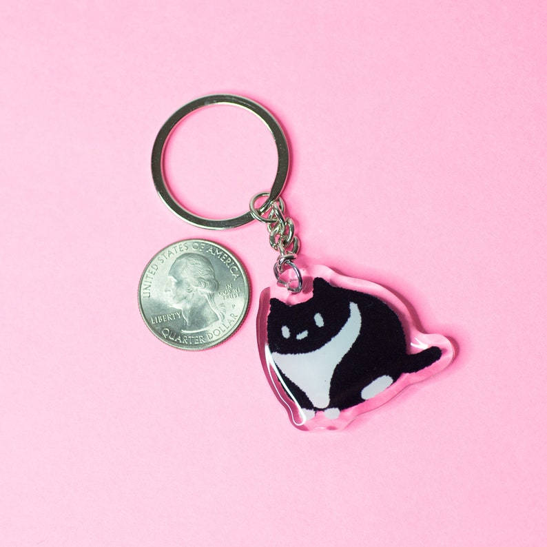 K&ouml;nnte beinhalten: Ein Schl&uuml;sselanh&auml;nger mit einer schwarzen und wei&szlig;en Katze und einem silbernen Schl&uuml;sselring. Die Katze sitzt auf einem durchsichtigen Acryl-Anh&auml;nger. Der Anh&auml;nger ist etwa so gro&szlig; wie eine 25-Cent-M&uuml;nze.
