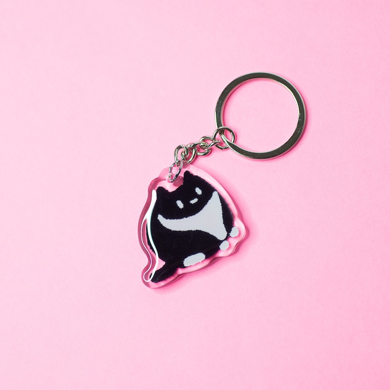 K&ouml;nnte beinhalten: Ein durchsichtiger Acryl-Schl&uuml;sselanh&auml;nger mit einem schwarz-wei&szlig;en Cartoon-Katzen-Design. Die Katze sitzt mit den Pfoten unter dem K&ouml;rper.