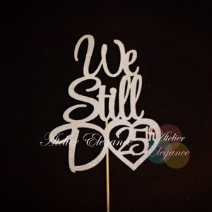 May include: A silver glitter cake topper with the words "We Still Do" and a heart with the number "25" inside. The topper is attached to a wooden stick, ideal for a 25th wedding anniversary.