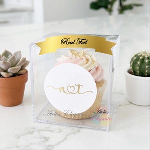 May include: A clear square plastic cupcake box featuring a gold foil banner at the top with the text Real Foil. A white circular sticker on the front displays the gold letters n and t connected by a heart. The box contains a cupcake with pink and white frosting.