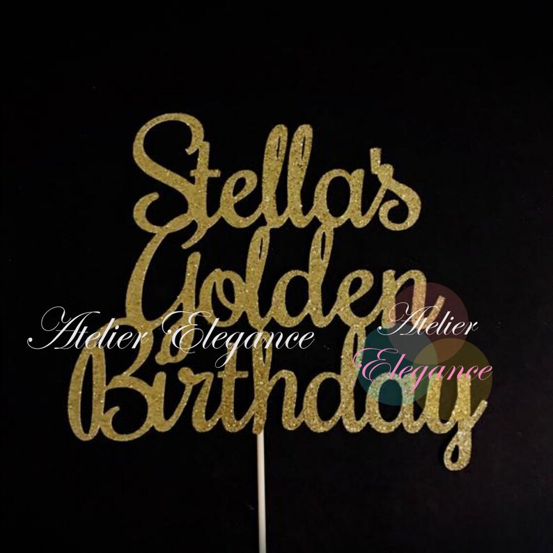 May include: A gold glitter cake topper with the words "Stella's Golden Birthday" in a cursive font. The topper is on a white stick, perfect for a birthday cake. The background is black.