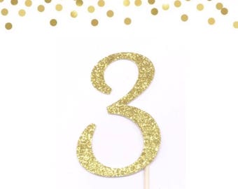 May include: Gold glitter number three cake topper on a white background. The number is on a wooden stick.