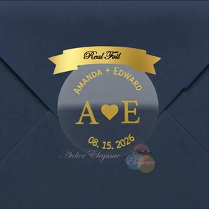 May include: A clear, round sticker with gold foil text and a gold ribbon. The sticker says "Real Foil" and features the names "Amanda + Edward", the initials "A ♥ E", and the date "08.15.2026". The sticker is on a navy blue envelope.