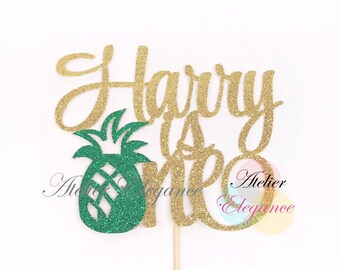 May include: Gold glitter cake topper with the text "Harry is one" and a green glitter pineapple. The topper is on a wooden stick.
