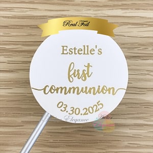 Peut inclure: Un cake topper rond blanc avec du texte doré. Il est écrit "Estelle's first communion" et la date "03.30.2025". Une bannière dorée au-dessus indique "Real Foil".