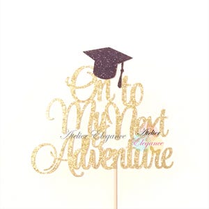 May include: A gold glitter graduation cake topper with the words "On to My Next Adventure" in a cursive font. A small, dark purple graduation cap sits above the text. The topper is attached to a wooden stick.