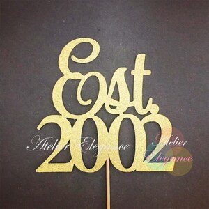 Puede incluir: Topper para pastel de purpurina dorada con el texto "Est 2002" en una fuente cursiva. El topper está en un palo de madera.