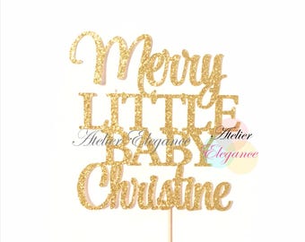 May include: A gold glitter cake topper with the words "Merry Little Baby Christine" in a script font. The topper is attached to a wooden stick for easy insertion into a cake. The background is white.