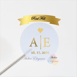 May include: A round white sticker with gold foil lettering and a gold heart. The sticker has the initials "A E" and the date "08.15.2026". A gold banner above reads "Real Foil".