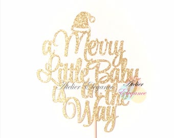 May include: A gold glitter cake topper with the phrase "a Merry Little Baby on the Way" and a Santa hat. The topper is on a wooden stick, ideal for a baby shower or Christmas celebration.