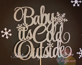 May include: Silver glitter cake topper with the text "Baby it's Cold Outside" and snowflakes. The topper is on a wooden stick.
