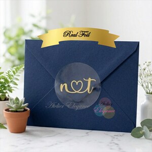 May include: A dark blue envelope sealed with a round transparent sticker featuring the gold foil letters n and t connected by a heart. A gold banner at the top displays the text Real Foil. The brand name Atelier Elegance is printed below.