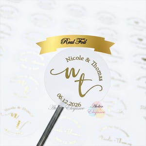 May include: A white round sticker with gold foil text. The sticker reads "Nicole & Thomas" with the initials "wt" and the date "06.12.2026". A gold banner above reads "Real Foil".