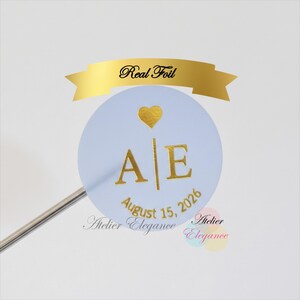 May include: A pale blue round sticker with gold foil details. The sticker displays the initials "A E", a gold heart, and the date "August 15, 2026". A gold banner above reads "Real Foil". The sticker is attached to a silver stick.