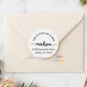May include: A circular white sticker with black text reading THE FUTURE MR & MRS erickson 12345 Sunnyside Drive Dallas, TX 70333, centred on a cream-coloured envelope. The sticker features a decorative script font for the name and serif font for the address.
