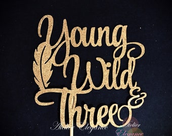 May include: Gold glitter cake topper with the words "Young Wild & Three" and a feather design.
