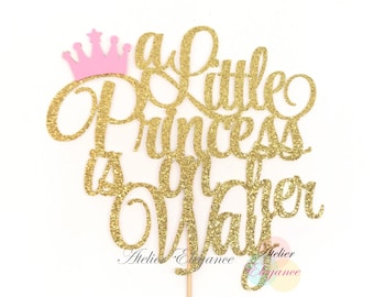 Könnte beinhalten: Goldener Glitzer-Kuchentopper mit dem Text "a little princess is on the way" und einer rosa Krone.