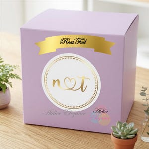 May include: A square lavender gift box featuring a gold foil banner with the text Real Foil. A circular white label with a gold dotted border displays the letters n and t connected by a heart. The text Atelier Elegance appears below the label.