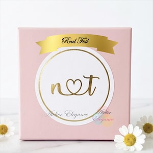 May include: A square light pink gift box featuring a white circular sticker with a gold foil border. The sticker displays the gold foil text "Real Foil" on a banner above the letters "n" and "t" connected by a heart. The text "Atelier Elegance" is printed below.