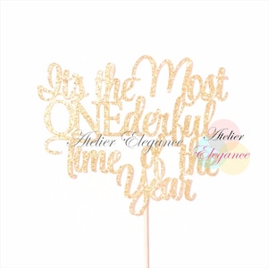 May include: A gold glitter cake topper with the phrase "It's the Most Wonderful Time of the Year" in a cursive font. The topper is attached to a thin, pink stick for insertion into a cake.