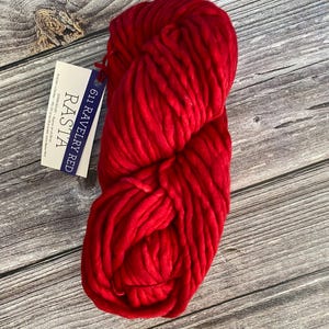 Puede incluir: Un ovillo de lana roja con la etiqueta "611 RAVELRY RED RASTIA" sobre una superficie de madera.