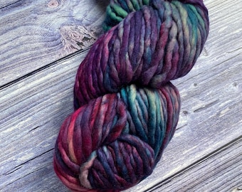 Könnte beinhalten: Ein Strang aus dickem, grobem Garn mit einer bunten Farbpalette aus tiefem Violett, Magenta, Blaugrün und Dunkelblau. Das Garn ist zu einer Acht gedreht und enthält ein kleines, weißes Papieretikett am oberen Abschnitt.