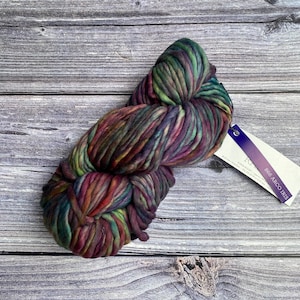 May include: A skein of colorful yarn in shades of green, purple, red, and brown. The yarn is thick and chunky, with a textured appearance. A white tag with the text "868 ARCO IRIS" is attached to the yarn.