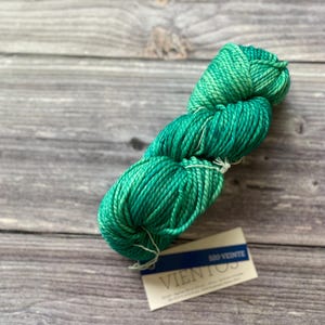 May include: A skein of vibrant green yarn with varying shades, resting on a wooden surface. The yarn is labeled "Vientos" with additional text. The color transitions from a darker teal to a lighter, brighter green.