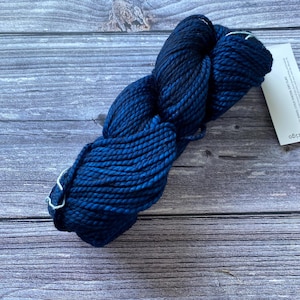 May include: A skein of dark blue yarn with lighter blue highlights. The yarn is thick and twisted, with a tag attached. The yarn is on a wooden surface.