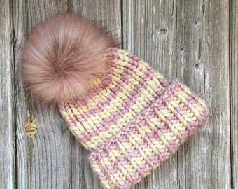Puede incluir: Un gorro de punto rosa y crema con un pompón rosa esponjoso. El gorro está hecho con un hilo grueso y tiene un patrón de chevron.
