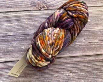 Könnte beinhalten: Ein Strang aus dickem, voluminösem Garn mit einer bunten Farbpalette aus tiefem Violett, gebranntem Orange, Goldgelb und Creme. Das Garn hat eine gedrehte, mehrfädige Textur und enthält ein kleines rechteckiges Papieretikett mit dem Text malabrigo.