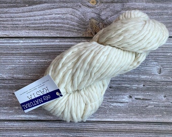 Könnte beinhalten: Ein Knäuel weißer Wolle mit der Aufschrift "063 NATURAL RASTIA" auf einer Holzoberfläche.