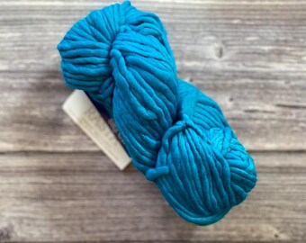 May include: A large skein of vibrant turquoise yarn. The thick, chunky yarn is tightly wound and has a soft, textured appearance. A small white tag is attached to the yarn. The yarn is displayed on a weathered, gray wooden surface.