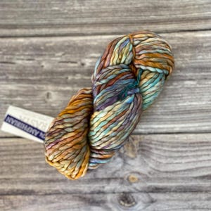 May include: A skein of yarn in shades of brown, blue, and green. The yarn is twisted into a ball shape and is sitting on a wooden surface. The label reads "The American Yarn"