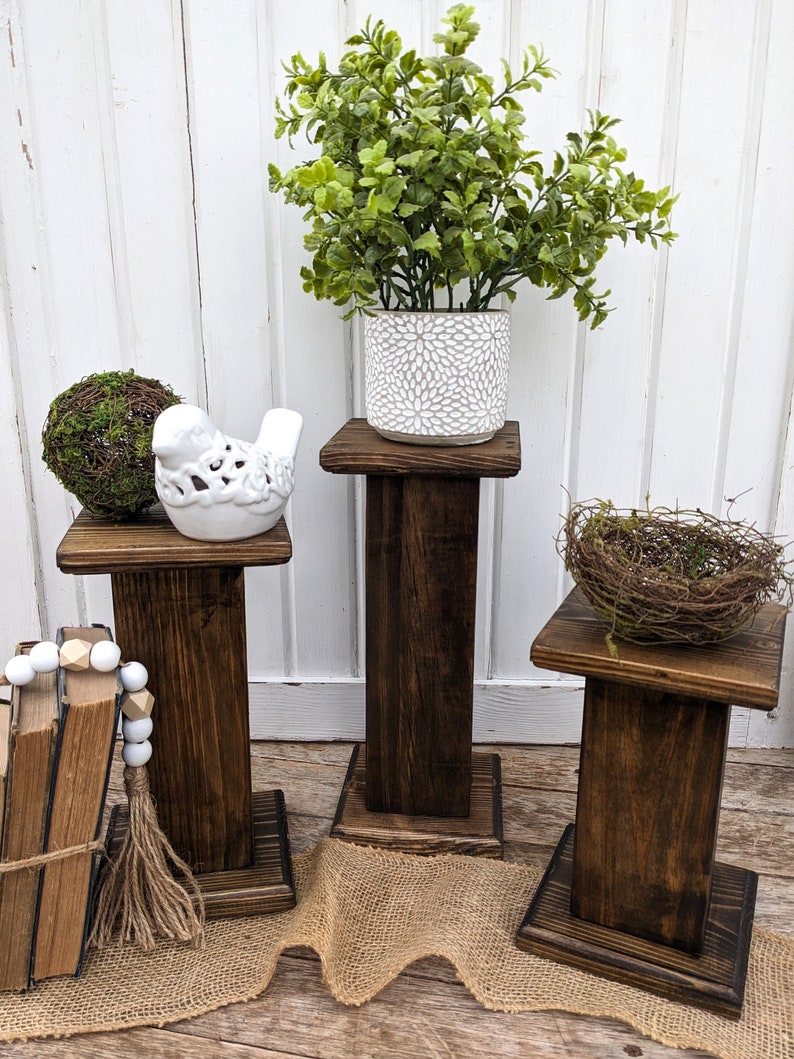 May include: Three dark stained wooden plant stands of varying heights. One holds a green plant in a white patterned pot, another a moss ball, and the third a bird's nest. A white bird figurine and a stack of books are also visible.