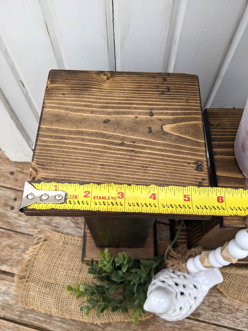 May include: A stained wooden riser with a square top, approximately 6 inches wide, and a dark brown finish. The wood grain is visible. A yellow Stanley measuring tape is placed on the top. The riser is part of a decorative display.