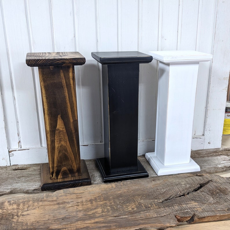 May include: Three wooden pedestals in varying colors: dark brown, black, and white. Each pedestal has a square top and base, with a rectangular column connecting them. The dark brown pedestal has a slightly angled column. The pedestals are displayed on a wooden surface.