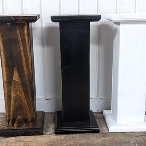 May include: Three wooden pedestals in varying colors: dark brown, black, and white. Each pedestal has a square base and a rectangular top, suitable for displaying items. The dark brown pedestal has visible wood grain, while the black and white ones have a smooth finish.