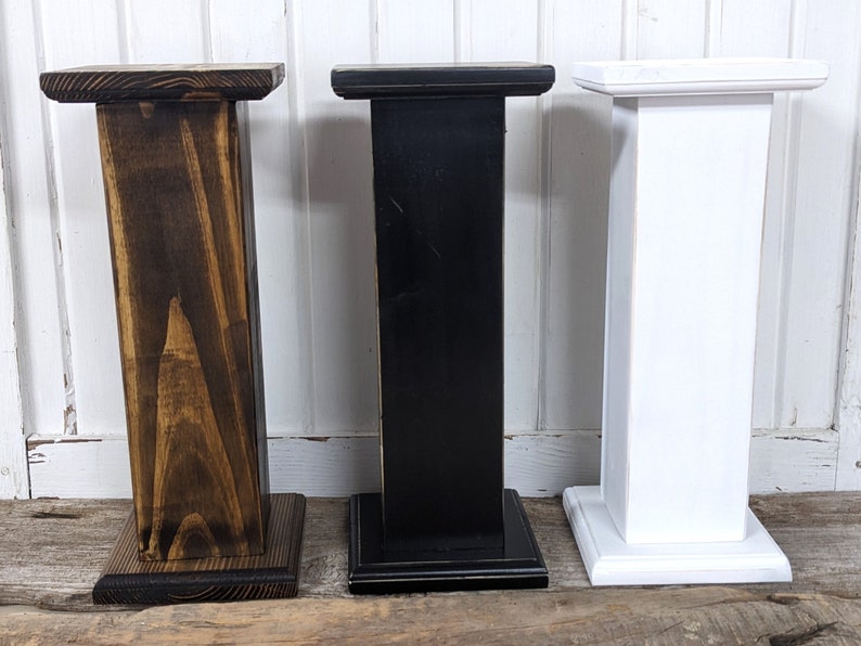 May include: Three wooden pedestals in varying colors: dark brown, black, and white. Each pedestal has a square base and a rectangular top, suitable for displaying items. The dark brown pedestal has visible wood grain, while the black and white ones have a smooth finish.