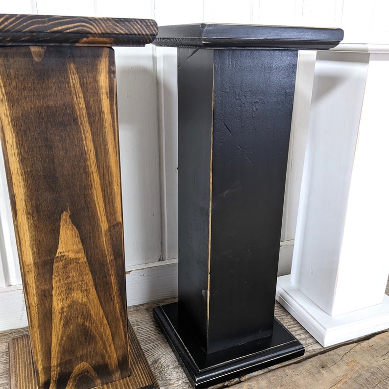 May include: Three wooden pedestals in varying colors: dark brown, black, and white. Each pedestal has a square top and base, with a rectangular column. The wood grain is visible on the brown pedestal.