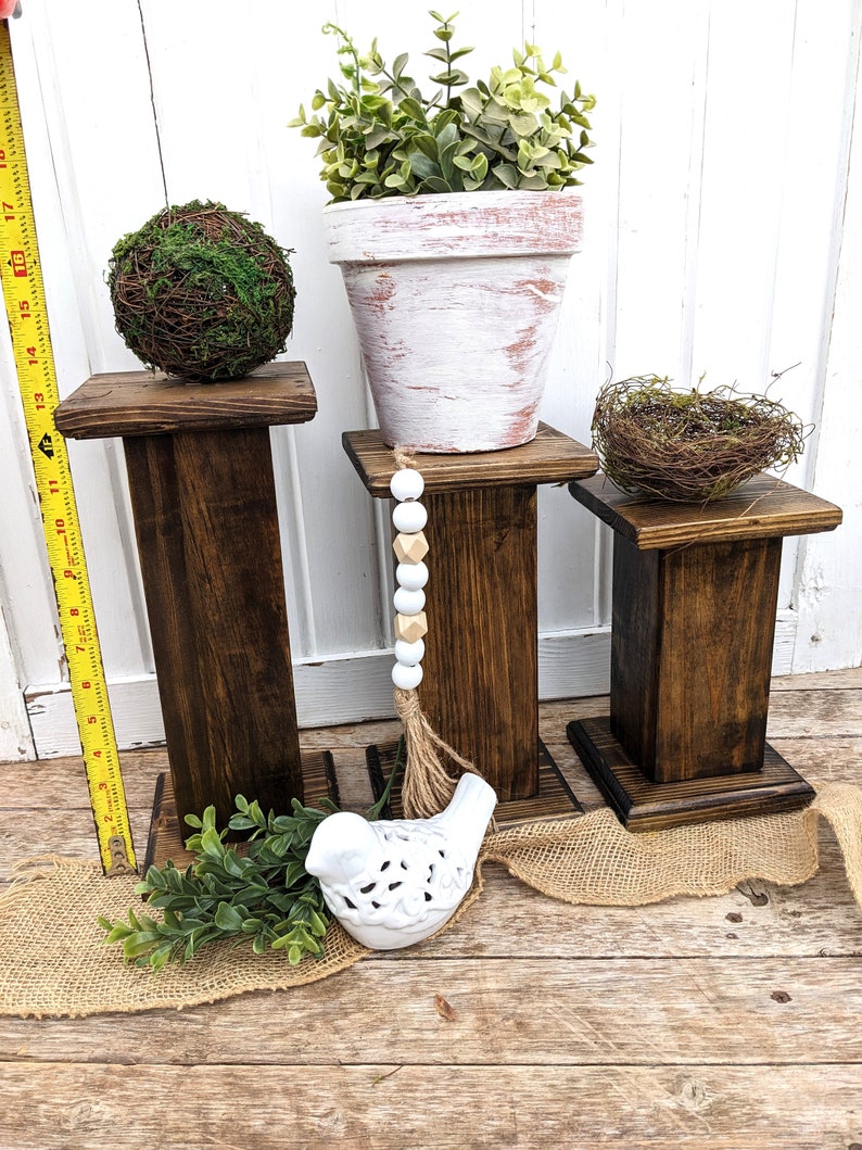 May include: Three dark stained wooden plant stands of varying heights. One stand holds a moss ball, another a white-washed terracotta pot with greenery, and the third a small bird's nest. A white ceramic bird and burlap accents complete the display.