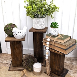 May include: Three dark stained wooden plant stands of varying heights. Each stand holds decorative items: a green plant in a white pot, a small succulent, a bird figurine, and books. A candle and decorative beads are also present.