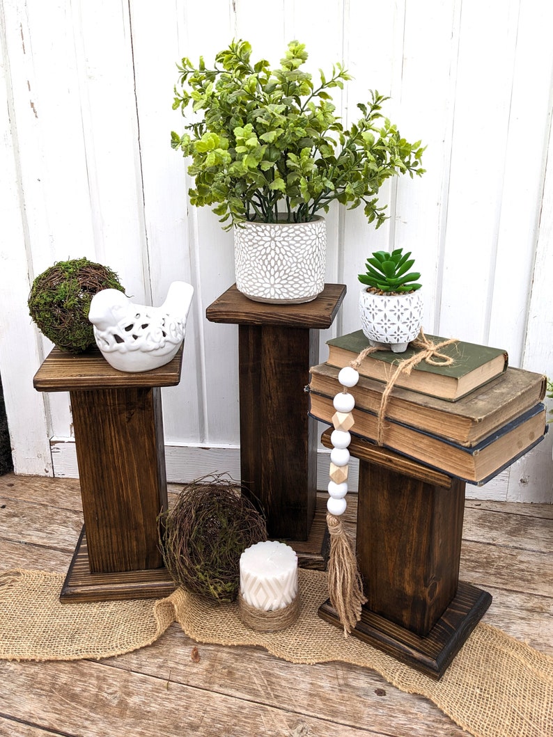 May include: Three dark stained wooden plant stands of varying heights. Each stand holds decorative items: a green plant in a white pot, a small succulent, a bird figurine, and books. A candle and decorative beads are also present.
