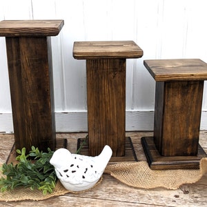 May include: Set of three wooden display stands in varying heights. Each stand features a square top and base, connected by a rectangular post. The wood has a dark brown stain, and the stands are arranged on a wooden surface with decorative elements.