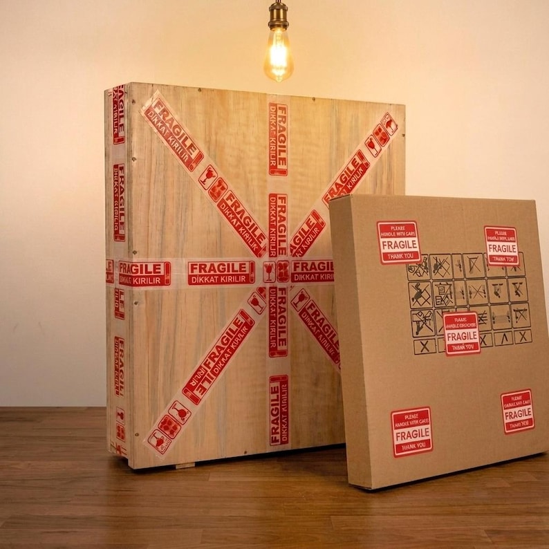 Puede incluir: Una gran caja de env&iacute;o rectangular de madera y una caja de cart&oacute;n cuadrada m&aacute;s peque&ntilde;a. La caja de madera est&aacute; envuelta en cinta roja y blanca con la palabra FRAGILE y DIKKAT KIRILIR. La caja de cart&oacute;n tiene pegatinas rojas de FRAGILE.