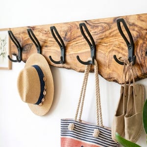 Può includere: Un appendiabiti da parete realizzato con una tavola di legno a bordo naturale e cinque ganci in metallo nero. Un cappello di paglia con una fascia blu navy, una borsa a righe con manici in corda e un paio di espadrillas in tela beige sono appesi.