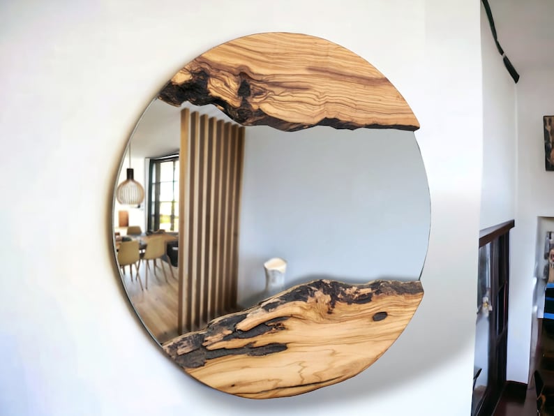 Puede incluir: Un espejo de pared circular que presenta dos secciones distintas de madera natural de borde vivo, con corteza oscura y texturizada y vetas de madera clara. Las piezas de madera est&aacute;n situadas en la parte superior e inferior, enmarcando el cristal.