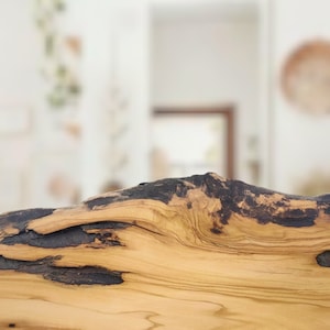 Puede incluir: Un trozo de madera de olivo natural con borde irregular, que presenta un patr&oacute;n de veta de color tostado claro contrastado por secciones oscuras y carbonizadas a lo largo del borde superior y un peque&ntilde;o nudo oscuro en la superficie.