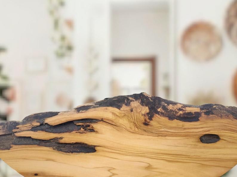 Puede incluir: Un trozo de madera de olivo natural con borde irregular, que presenta un patr&oacute;n de veta de color tostado claro contrastado por secciones oscuras y carbonizadas a lo largo del borde superior y un peque&ntilde;o nudo oscuro en la superficie.